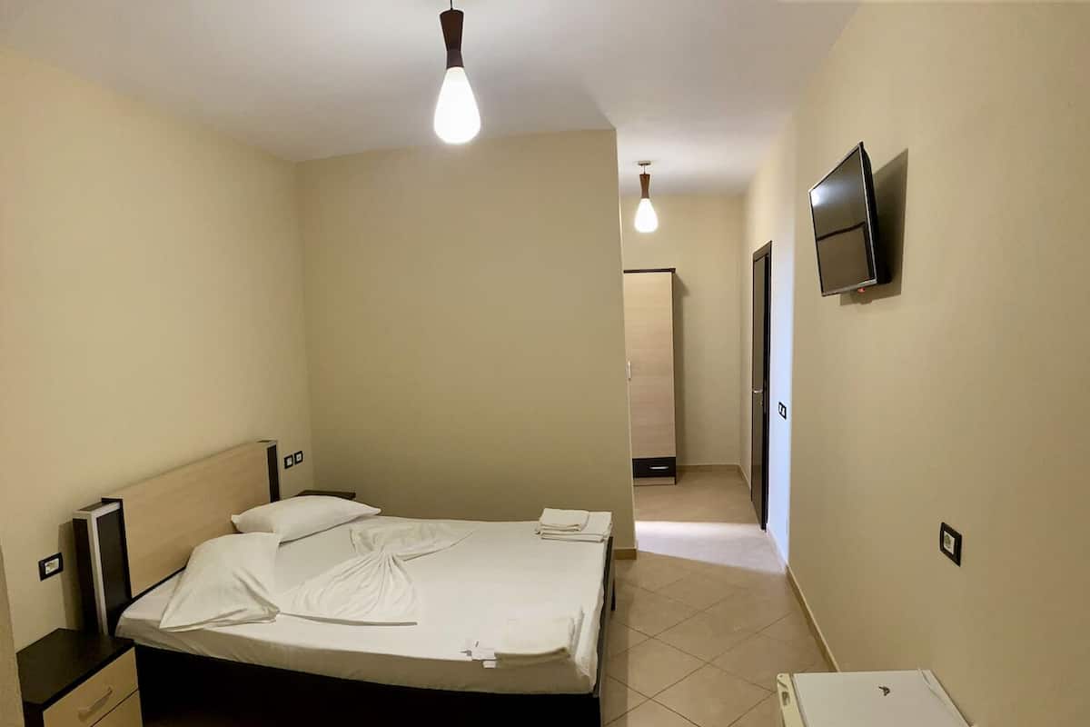Quarto de hotel com cama, mesas de cabeceira, TV fixada na parede e um pequeno armário branco.