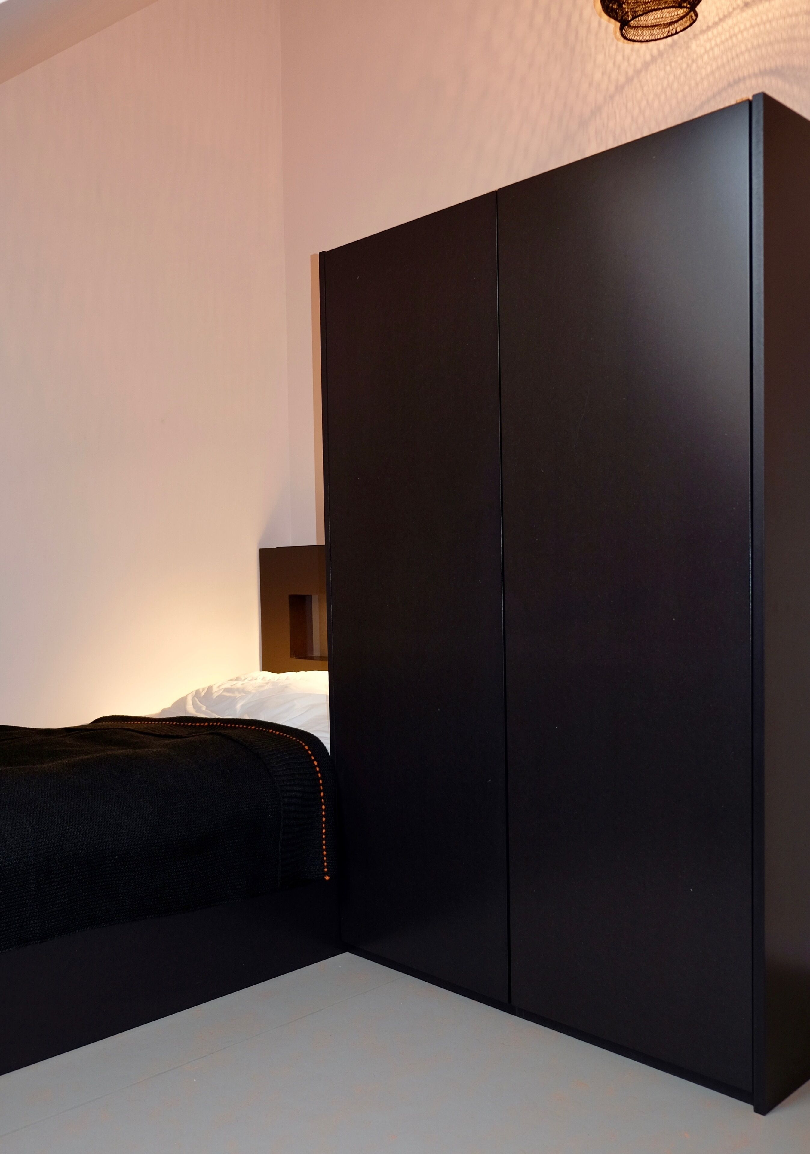 A modern bedroom with a large black wardrobe, a bed with a black cover, and a small bedside table with a lamp.