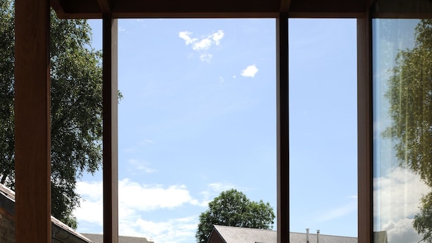 A view through a wooden-framed window of a green lawn, a gravel path, and a brick building with a sloped roof. - Le Presbytère (Engis)