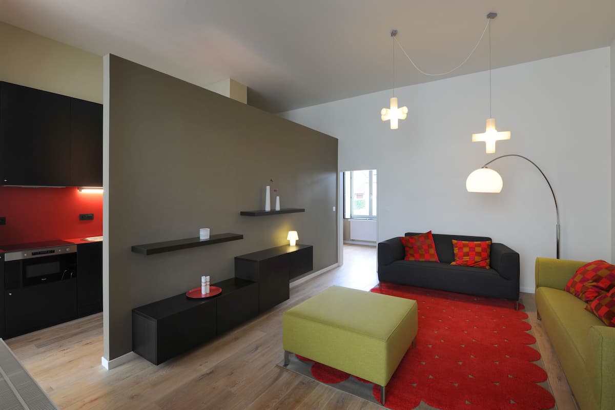 A modern living room with a sofa, ottoman, and a red rug. There is a kitchen area with a red-lit stove and black cabinets.