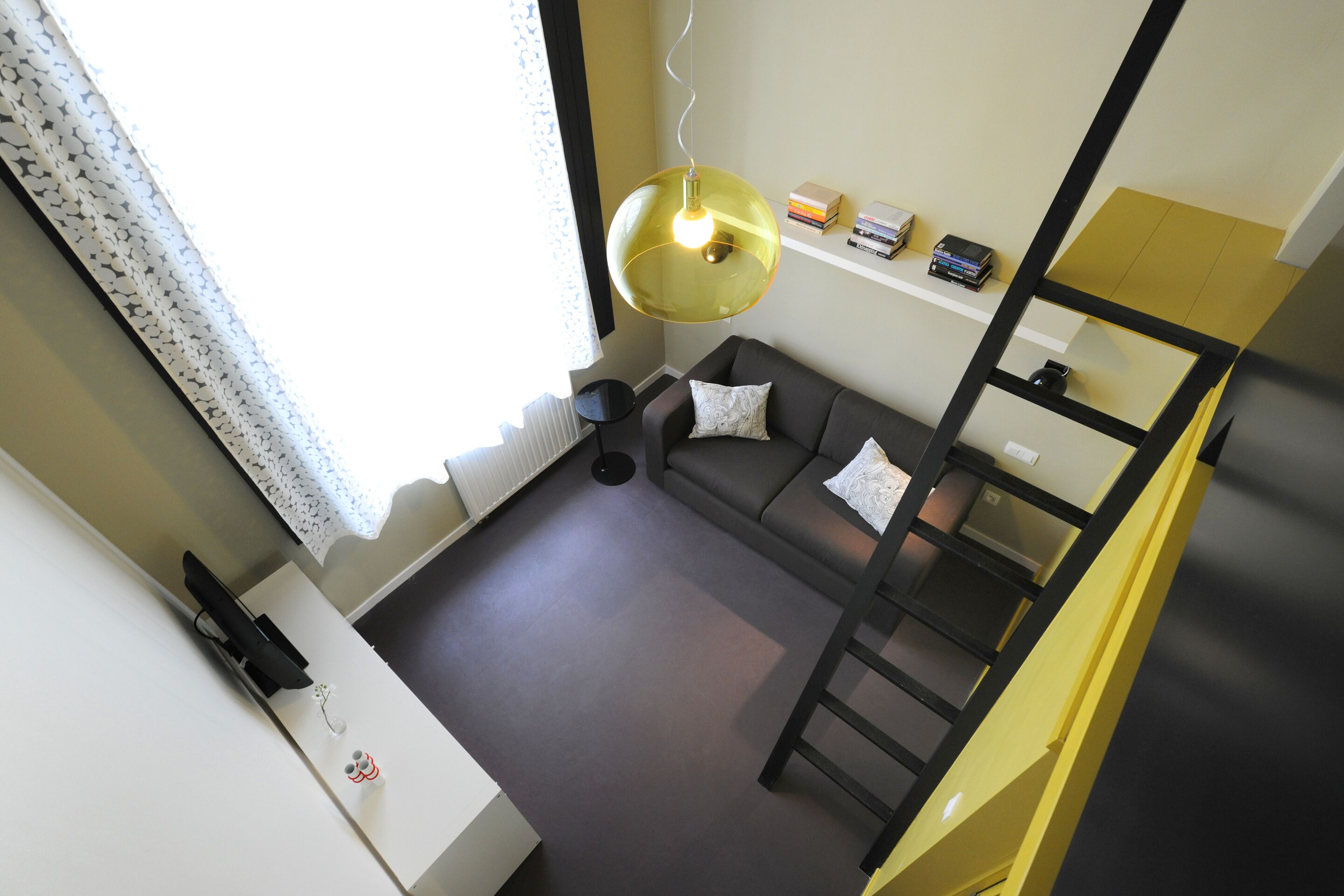 A compact room with a sofa, a small table, a hanging light fixture, and a shelf with books and a box.