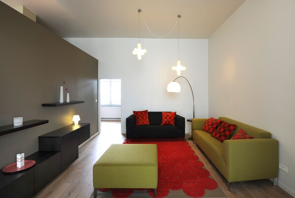 Una sala de estar moderna con un sofá negro, un taburete verde y una alfombra roja. Hay dos lámparas colgantes y una lámpara de pie.