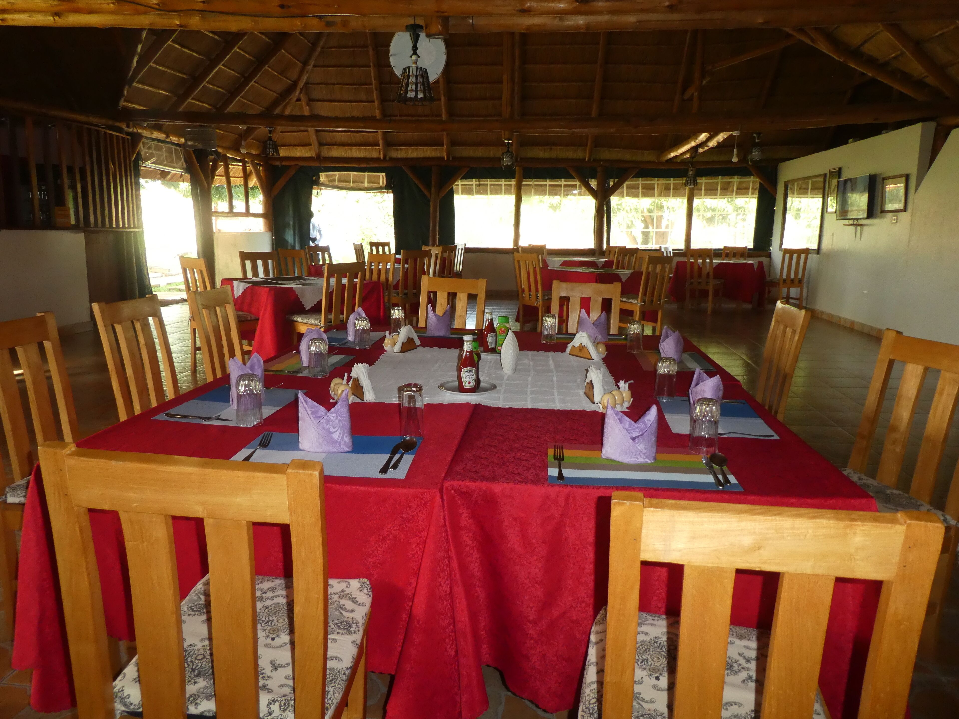 A dining area with tables set for multiple guests, wooden chairs, and a thatched roof structure.