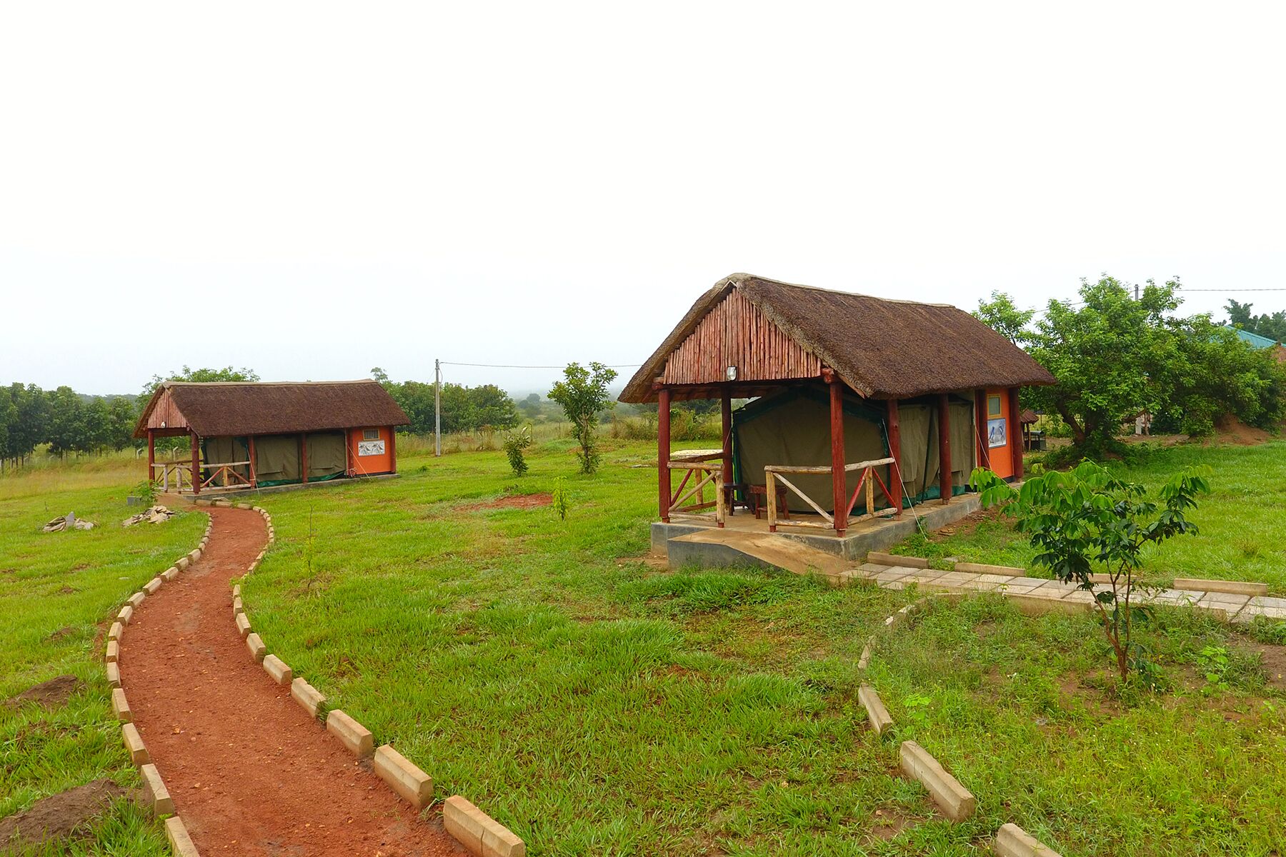 Un terrain de camping situé en pleine campagne, avec deux cabanes aux toits de chaume, un chemin de terre et une végétation luxuriante tout autour.