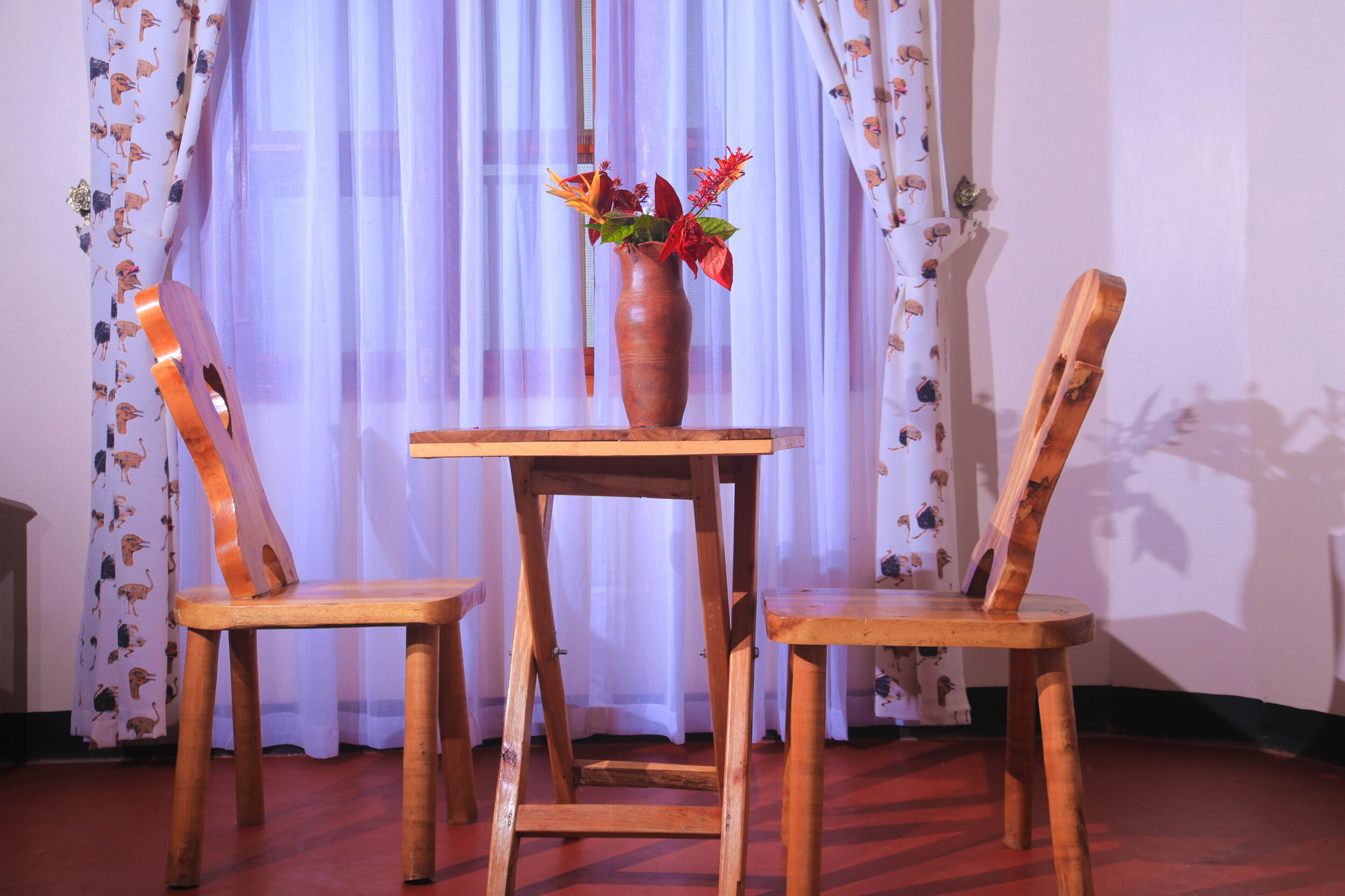 A room with a wooden table and two chairs, a vase with flowers, and a window with sheer curtains.