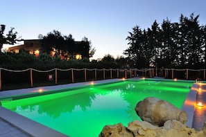 Une piscine éclairée de lumière verte, entourée d’une clôture et d’arbres.