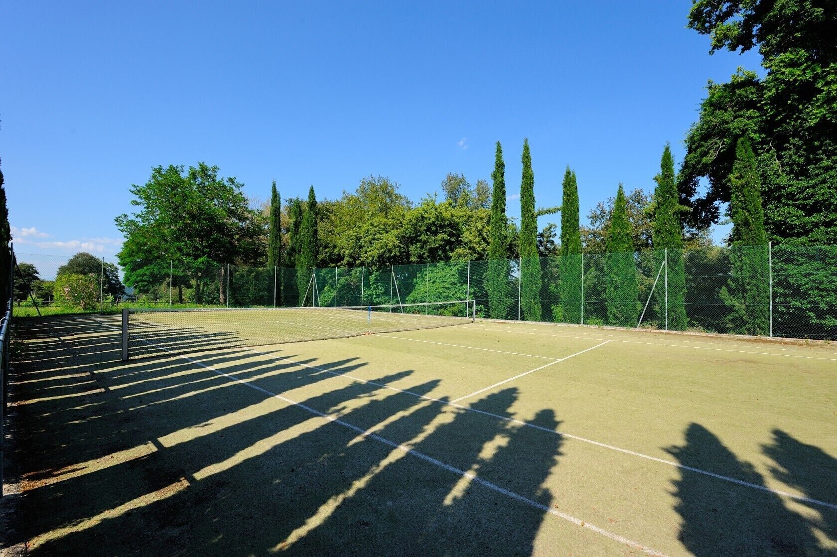An outdoor tennis court with a net, surrounded by trees and a fence.