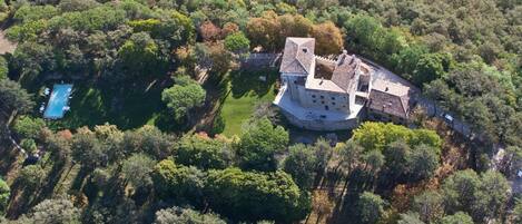 An aerial view of a castle-like structure surrounded by dense forest, with a swimming pool visible to the left.