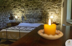 Een slaapkamer met een stenen muur, een bed met een metalen hoofdeinde, twee nachtkastjes met lampen, en een brandende kaars op een tafel.