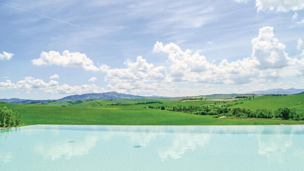 Piscina com vista para campos verdes e montanhas ao longe, sob um céu parcialmente nublado.