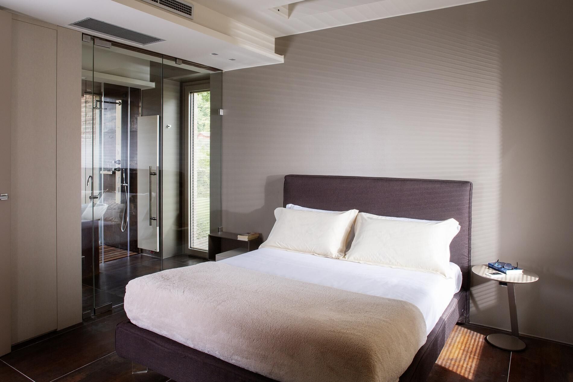 A modern hotel room with a large bed, a glass shower enclosure, and a small bedside table with a lamp.