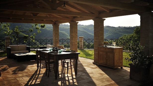 Une terrasse extérieure avec une table et des chaises en bois, une cheminée en pierre et une vue sur des collines ondoyantes.