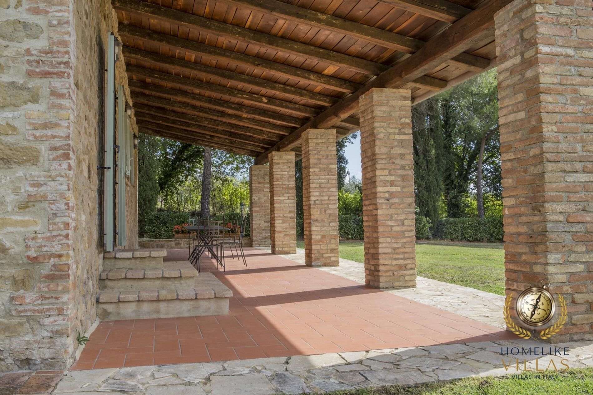 Un área de patio techada al aire libre, con piso de piedra y vigas de madera, rodeada de muros de ladrillo y vegetación.