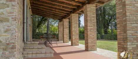 Un área de patio techada al aire libre, con piso de piedra y vigas de madera, rodeada de muros de ladrillo y vegetación.