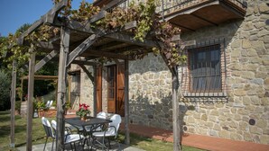 Ein Steingebäude mit einer Weinbepflanzung an einer Pergola, eine Steinterrasse mit Tisch und Stühlen und eine Steinsäule mit einem Fenster.