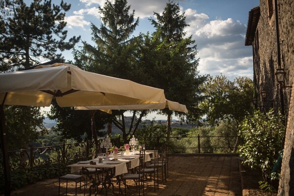 An outdoor dining area with a table set for four under a large umbrella, surrounded by trees and a stone wall. - Villa Greta (Orvieto)