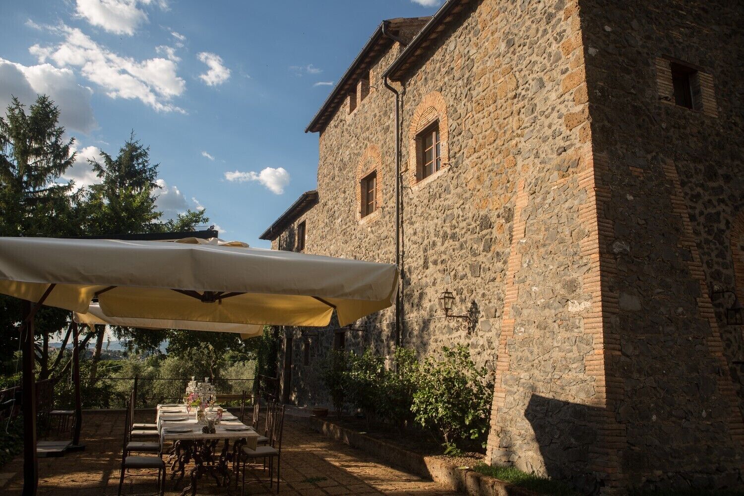 Un bâtiment en pierre avec une table et des chaises placées sous un grand parasol, le tout entouré d’arbres.