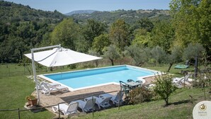 A rectangular swimming pool with a white umbrella, surrounded by white lounge chairs and a table. The pool is set in a grassy area with a view of rolling hills and trees in the background. - Villa Claudia (Gualdo)