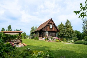 Une maison rustique en bois, dotée d’un toit à pente, entourée d’un jardin bien entretenu où poussent diverses plantes et fleurs.