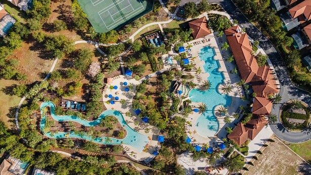 Vista aérea de un complejo turístico con una amplia piscina, varias zonas de descanso y una cancha de tenis.