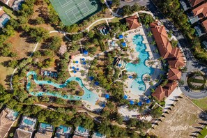 An aerial view of a resort with a large swimming pool, multiple lounge areas, and a tennis court.