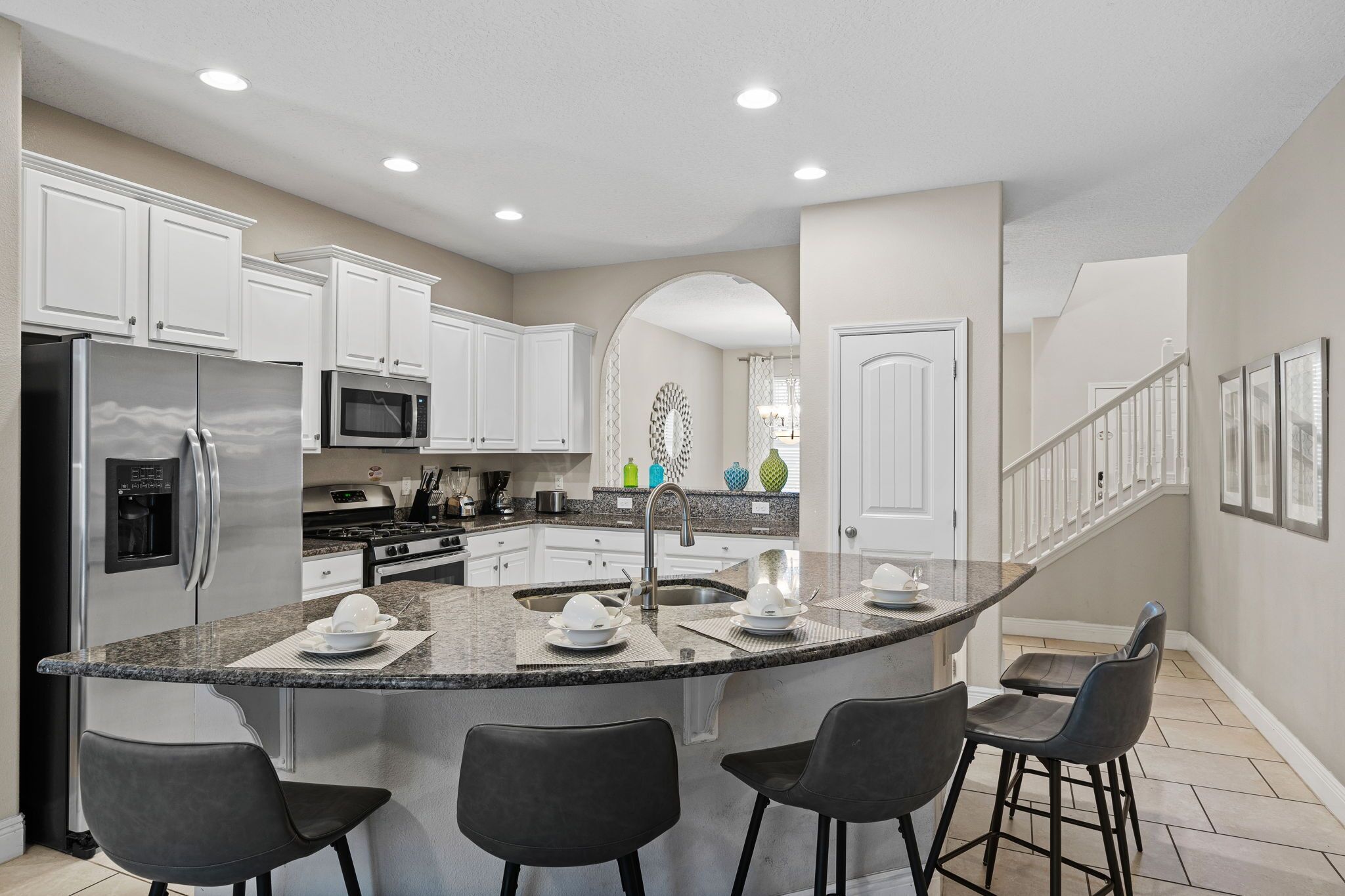A modern kitchen with white cabinets, a stainless steel refrigerator, a microwave, and a granite countertop island with bar stools.