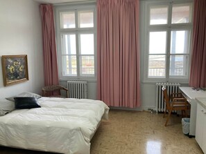 A bedroom with a bed, two windows with curtains, a radiator, a chair, a desk, and a picture on the wall.
