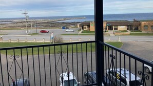 A view from a balcony overlooking a parking lot with cars, a building with a green roof, and a body of water in the background. - The Couvent (Sainte-Anne-des-Monts)