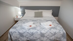 Ein ordentlich bezogenes Bett mit einer grauen, gemusterten Tagesdecke, zwei aufgerollte weiße Handtücher mit roten Blumen und ein graues Kopfkissen.