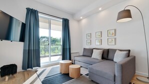 Een moderne woonkamer met een grijze bank, twee ronde houten bijzettafeltjes, een zwart pluchen pouf, een flatscreen televisie en een groot raam met blauwe gordijnen.
