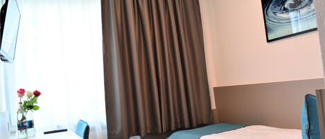 A hotel room with a desk, a bed with a blue bedspread, a large window with curtains, and a painting on the wall.
