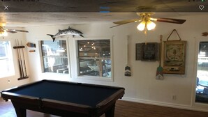A pool table in a room with wooden floors, a ceiling fan, and a large window with a view of boats outside.