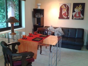 Uma área de refeições com mesa de vidro, cadeiras e toalha de mesa vermelha. Há um sofá preto e uma estante de livros ao fundo.