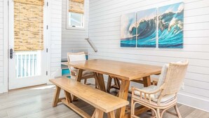 A dining area with a wooden table and benches, white paneled walls, and a large wave painting. - Big Wave | Private Pool Optional Heat, Hot Tub (Tybee Island)