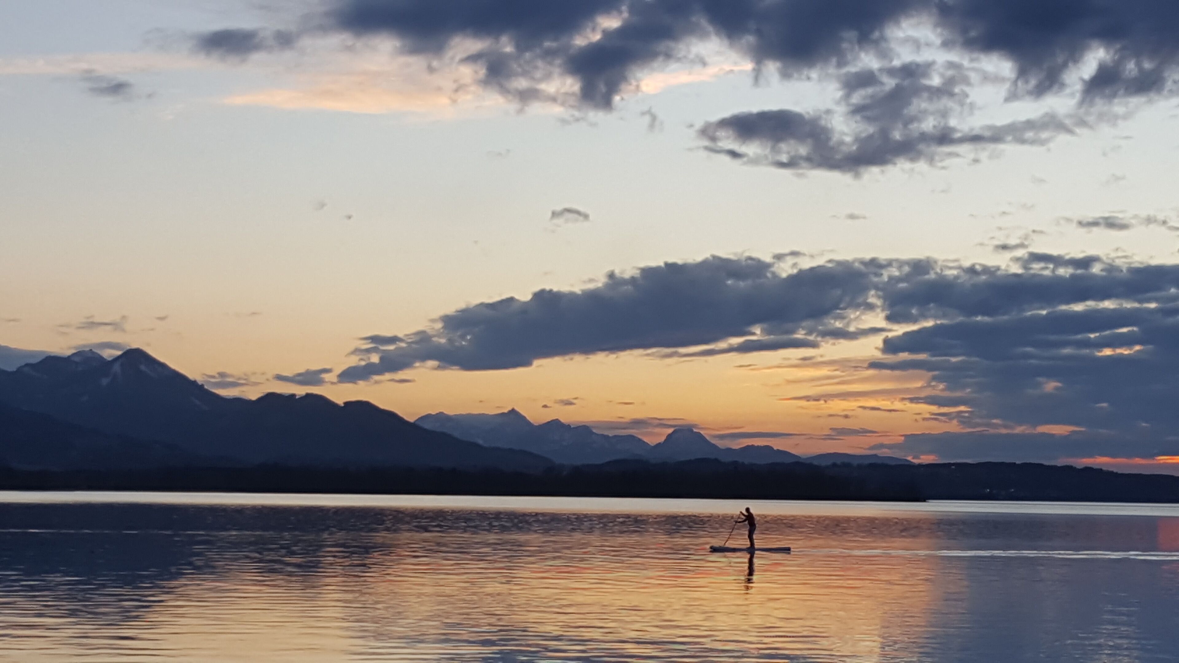 Un lago tranquilo con una persona haciendo paddleboard, montañas al fondo y un cielo al atardecer.