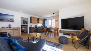 Ein modernes Wohnzimmer mit einer Couch, einem Couchtisch und einem Fernseher. Es gibt eine Küchenzeile mit Schränken und einem Esstisch.