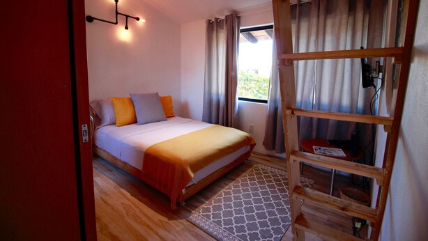 Quarto com cama, escada de madeira, janela com cortinas e luminária fixada na parede.