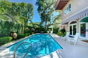 A swimming pool with a deck, surrounded by lush greenery and a house with a covered patio.
