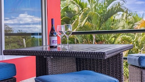 Une table en osier, avec une bouteille de vin et deux verres, sur un balcon dont le mur est peint en rouge et où l’on aperçoit des palmiers au fond.