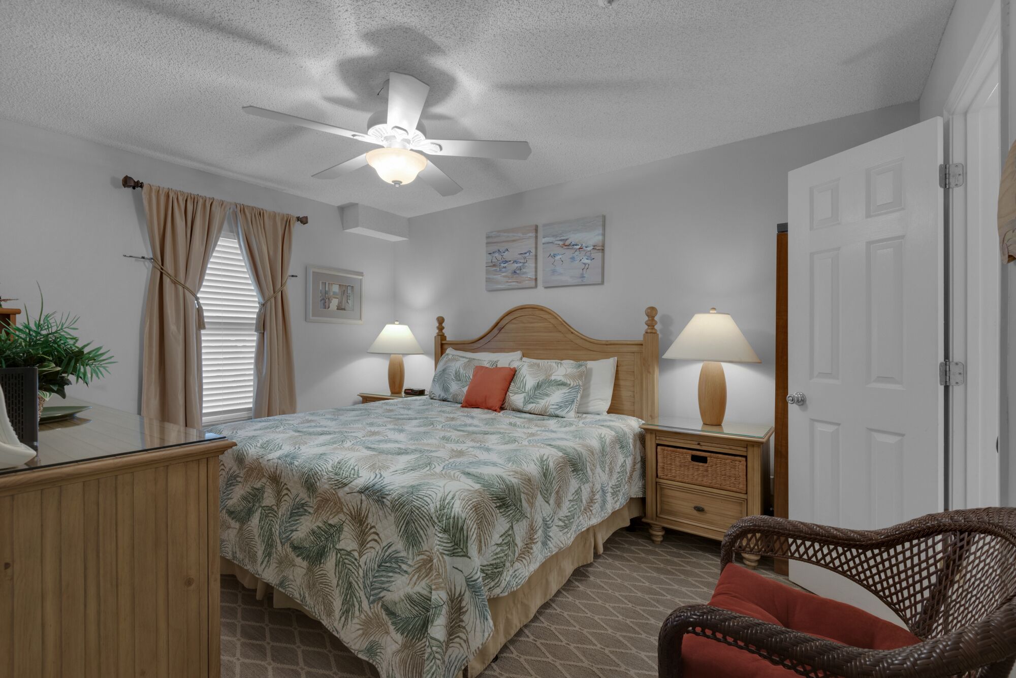 A bedroom with a bed, two bedside tables with lamps, a ceiling fan, a door, and a wicker chair.