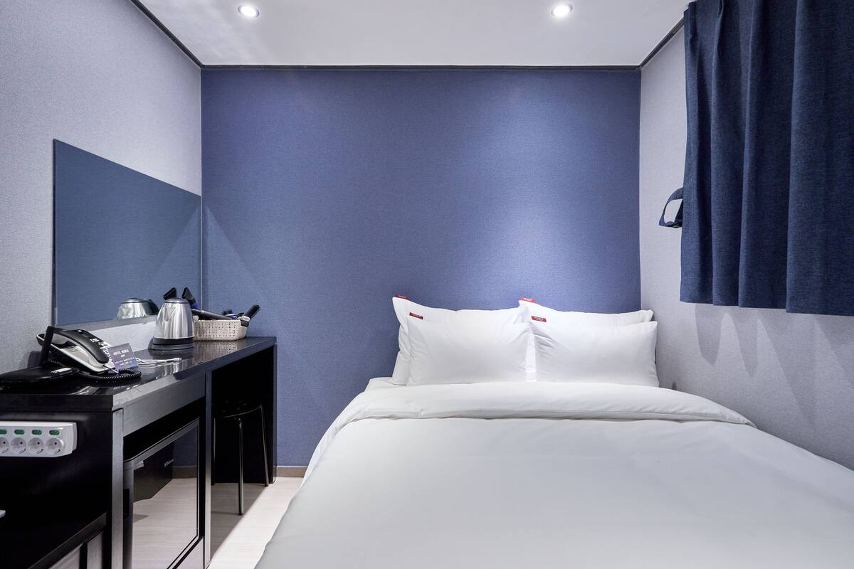 Una habitación de hotel con cama, escritorio, teléfono y una cesta, además de un espejo montado en la pared.