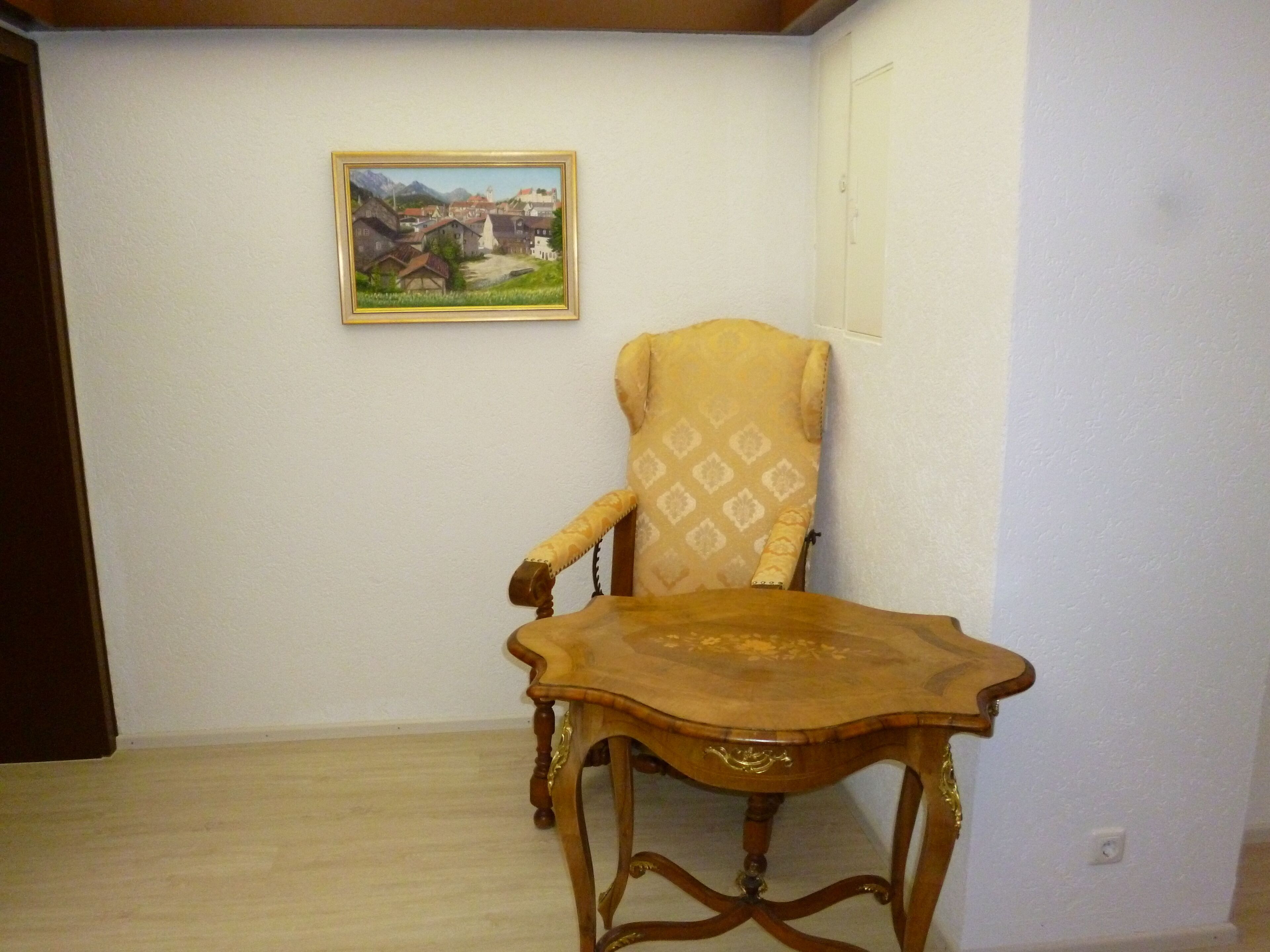 A room with a wooden chair and a table, a framed painting on the wall, and a door.