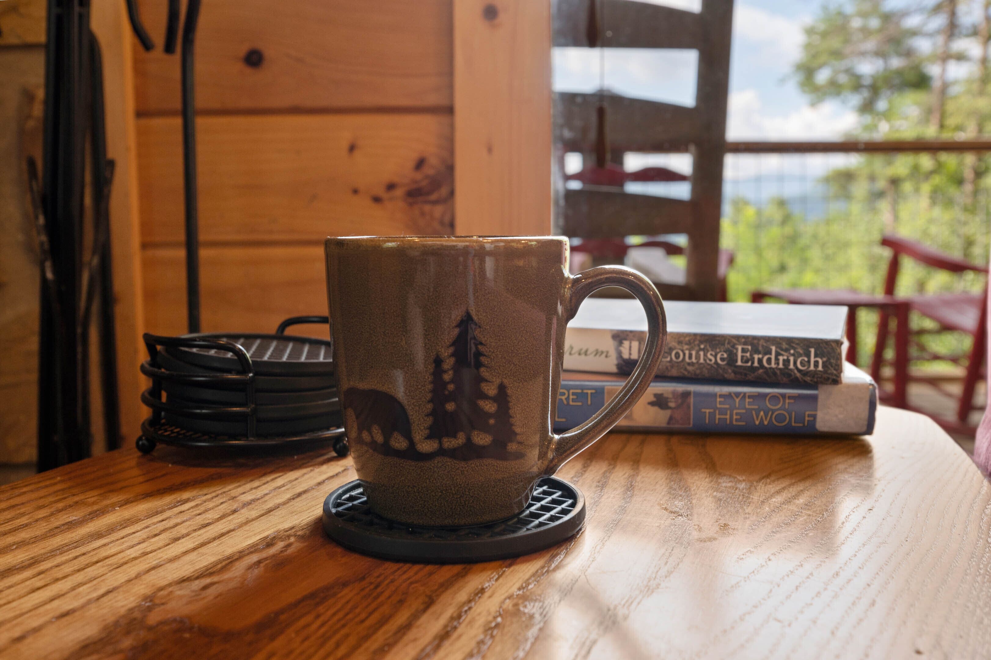 A wooden table with a coaster mug featuring a forest design, a stack of books, and a view of the outdoors.