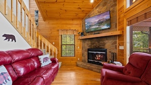 A log cabin interior with a stone fireplace, a red leather sofa, and a television mounted above the fireplace. - Hot Tub & Pool Table + Fire Pit - Secluded (Sevierville)