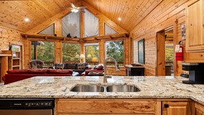 Interior de una cabaña de madera con cocina, comedor y un amplio ventanal con vistas a la vegetación.