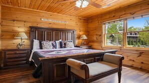 A wooden cabin bedroom with a bed, two bedside tables, a chair, a window with a view, and a ceiling fan. - Splash Mountain Magic- Views, Indoor pool, Game Room w/ Pool Table & Arcade (Sevierville)