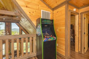 A wooden cabin with a wooden deck, a Multicade arcade machine, and a view of the mountains through the windows.
