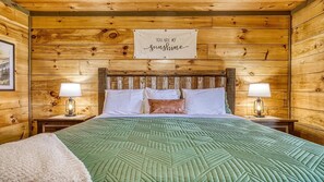 A wooden cabin room with a bed, two bedside lamps, a framed sign above the bed, and a green patterned bedspread. - The Appalachian- Mtn Views, Private/ Heated Indoor Pool, Game Room, Hot tub (Sevierville)
