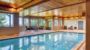 Une piscine intérieure avec des poutres en bois au plafond, un espace détente avec des fauteuils et de grandes fenêtres.