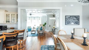 Une cuisine spacieuse avec des meubles blancs, un coin repas avec une table et des chaises en bois, et un salon avec des fauteuils bleus et un lustre.